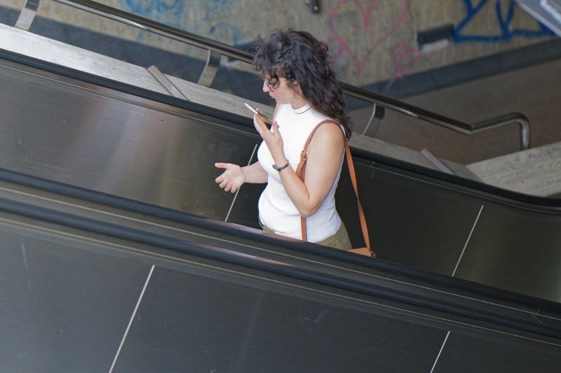 6 - Person with dark, curly hair wearing a white sleeveless top and a brown crossbody bag while standing on an escalator
